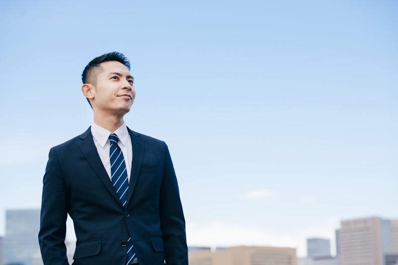 晴天下、斜め上を見上げて自身ありげに微笑む、スーツ姿で刈り上げショートカットの男性